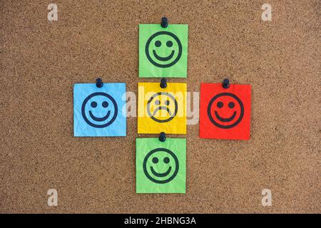 Una nota di carta con un volto triste con volti felici intorno ad esso su un bordo del corkboard. Concetto di rifiuto sociale. Primo piano. Foto Stock