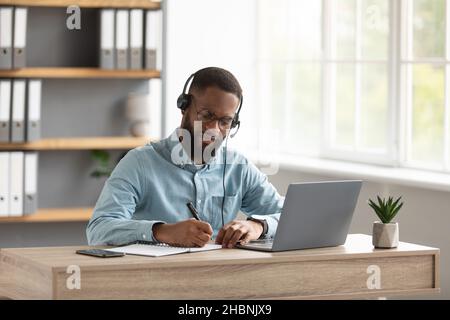 Serio impegnato millenario africano americano barbuto ragazzo manager in occhiali, cuffie lavoro o studio con notebook Foto Stock
