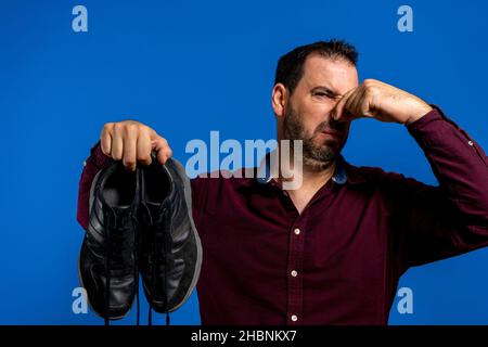 Uomo bearded vestito in una camicia viola che tiene le sue sneakers con un volto disgustato a causa del cattivo odore che danno fuori, isolato su sfondo blu Foto Stock