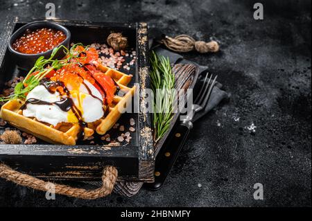 Delizioso waffle con filetto di salmone affumicato, uova in camicia e caviale rosso. Sfondo nero. Vista dall'alto. Spazio di copia Foto Stock