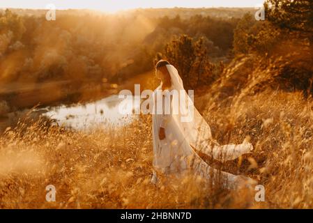 Foto di una sposa sognante in un abito lussureggiante vortice vicino al lago al tramonto Foto Stock