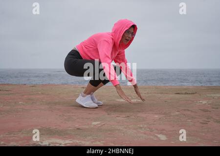 Donna con maschera esegue squat di fronte al mare. Nuova normalità. Sport Foto Stock