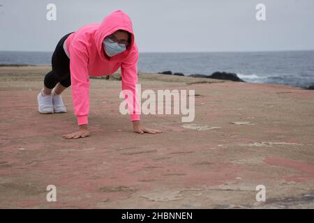 Donna mascherata esegue push-up di fronte al mare. Nuova normalità. Sport Foto Stock