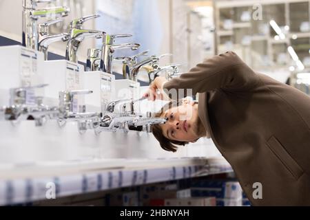 Un giovane uomo in un cappotto marrone sceglie con cura un miscelatore d'acqua in un supermercato di costruzione, le riparazioni idrauliche in un appartamento. Foto Stock