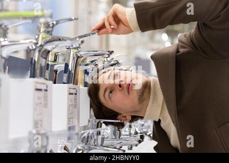 Un giovane uomo in un cappotto marrone sceglie con cura un miscelatore d'acqua in un supermercato di costruzione, le riparazioni idrauliche in un appartamento. Foto Stock
