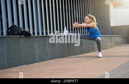 Scatto a tutta lunghezza di allegro medio vecchio sportswoman sorridente mentre riscalda il suo corpo, avendo workout all'aperto Foto Stock