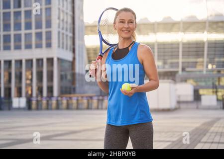 Allegro sportivo di mezza età sorridente, con racchetta da tennis e una palla pronta per giocare a tennis in città Foto Stock