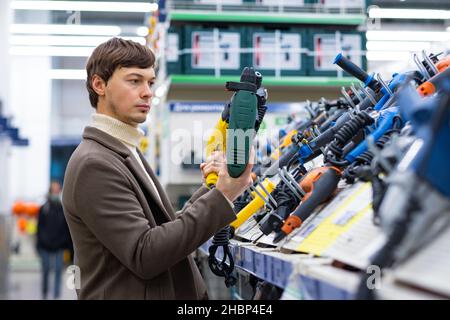 Un giovane uomo in un cappotto marrone seleziona con cura un perforatore in un supermercato per le riparazioni in un appartamento Foto Stock