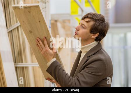 l'uomo riuscito in un cappotto marrone seleziona con cura le tavole di legno per la costruzione in un negozio di riparazione Foto Stock