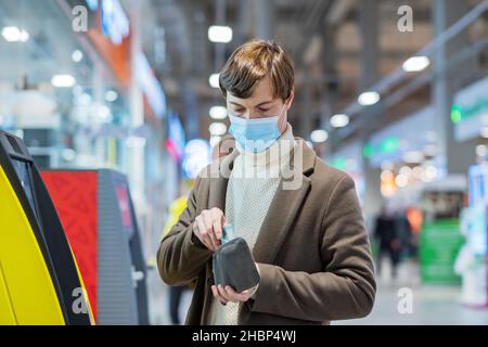 Un uomo riuscito in un cappotto marrone e maschera mette i soldi in un portafoglio ad un bancomat, prelevando i fondi da una carta bancaria Foto Stock