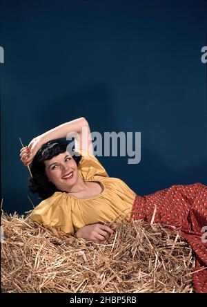 Dark haired teen girl in yellow blouse and red print skirt laying in pile of hay Foto Stock