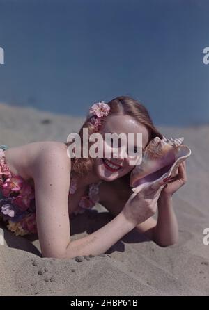 Giovane donna in un piano di balneazione fiorito, adagiato sulla spiaggia tenendo una grande conchiglia di mare fino al suo orecchio Foto Stock