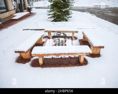 Quattro panchine di legno innevate si trovano intorno a un barbecue decorativo figurato in un'area coperta di neve Foto Stock