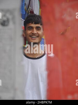 Fuoco selettivo di un giovane indiano felice che guarda la macchina fotografica mentre si trova intorno alle bandiere di preghiera buddiste Foto Stock