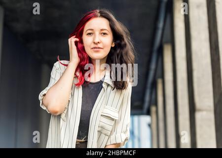 Ritratto lifestyle di donna elegante sorridente con capelli e bracciali colorati rosso e nero ondulati sui denti. Colore dei capelli a doppia tecnica Foto Stock