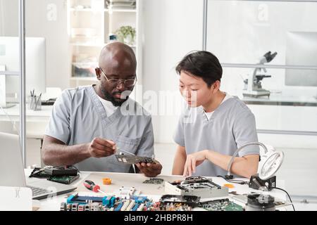 Giovane uomo africano con circuito stampato di consultare la sua collega femmina mentre spiega come risolvere i piccoli dettagli o microchip Foto Stock