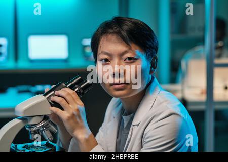 Giovane scienziata o tecnico fidato con microscopio che vi guarda mentre siete seduti sul posto di lavoro in laboratorio Foto Stock