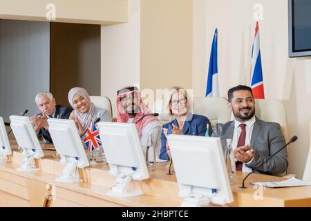 Felici delegati interculturali in abbigliamento formale che si aggrappano le mani all'oratore dopo il resoconto mentre si siede lungo il tavolo nella sala conferenze Foto Stock