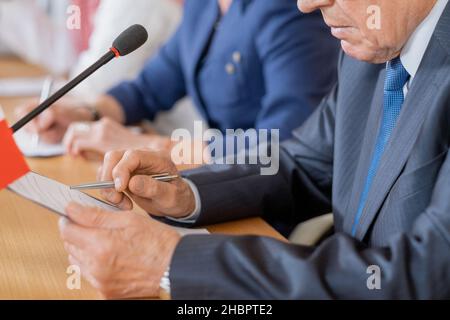 Mani di un delegato maturo che indicano il documento negli appunti mentre fanno rapporto per tabella contro fila di colleghi stranieri Foto Stock