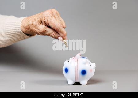 La mano rugosa di una donna anziana mette una moneta in euro in una banca piggy. Sfondo grigio. Il concetto di risparmio e bancario. Foto Stock