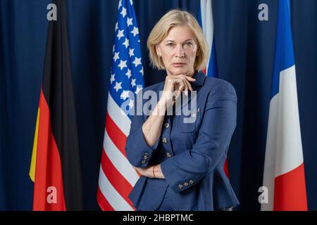 Delegata femminile matura contemporanea in abbigliamento formalwear che ti guarda mentre ti si levano contro le bandiere di diversi paesi Foto Stock