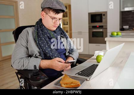 Giovane uomo in casualwear seduto in sedia a rotelle mentre scorre tra i contatti in smartphone di fronte al portatile Foto Stock