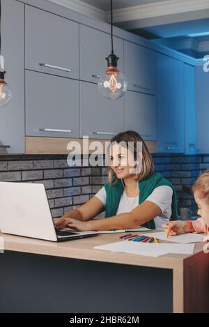 Giovane donna caucasica che lavora in linea sul laptop con la figlia che fa i suoi compiti in cucina. Rete Internet. Formazione online. Foto Stock