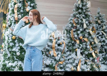 Ritratto di Capodanno di bella ragazza caucasica sorridente in lana accogliente caldo azzurro maglione in piedi vicino decorato albero di Natale. Persona felice Foto Stock