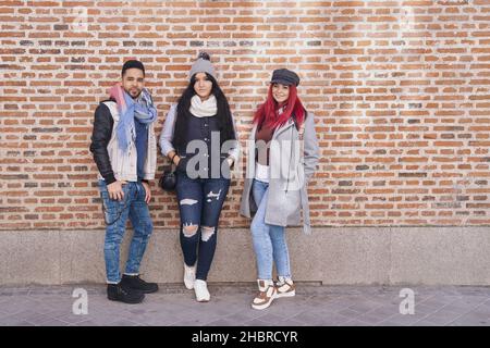 Positivo multirazziale giovani amici maschili e femminili indossando abiti casual trendy in piedi vicino a muro di mattoni e sorridendo alla macchina fotografica Foto Stock