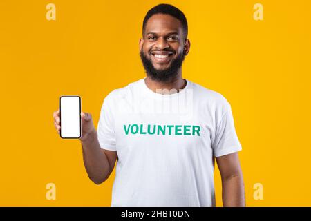 African Volunteer Man mostra lo schermo vuoto dello smartphone, sfondo giallo, Mockup Foto Stock