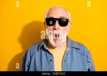 Ritratto fotografico di uomo anziano stupito con bocca aperta indossando giacca di jeans di vetro solare isolato su sfondo giallo brillante Foto Stock