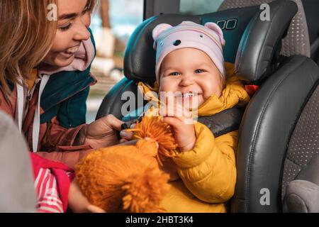 Giovane madre caucasica sorridente sta preparando il suo bambino felice per un viaggio in auto. Una donna allaccia le cinture di sicurezza in un seggiolino per bambini nello schienale. Chiudi u Foto Stock