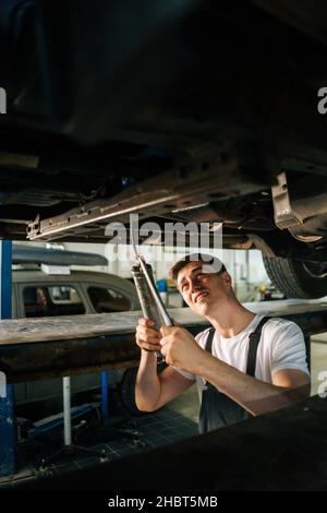 Ritratto verticale di serio bel meccanico professionale maschile auto in uniforme in piedi in buca di ispezione e lavorando con l'utensile. Foto Stock