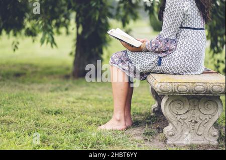 Un dettaglio di una donna solitaria seduta su una panca parco con un libro Foto Stock