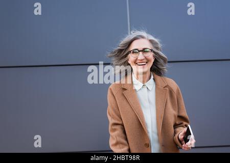 Donna d'affari matura in cappotto con smartphone con schermo vuoto e sorridente alla fotocamera all'aperto Foto Stock