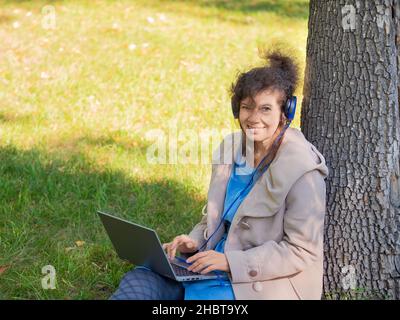 Felice sorridente donna di mezza età in cuffie con computer portatile nel parco della città. Foto Stock