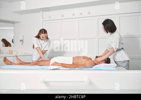 Le donne chiropratica in uniformi lavorano con pazienti maschi maturi sdraiati sul divano in un salone termale Foto Stock