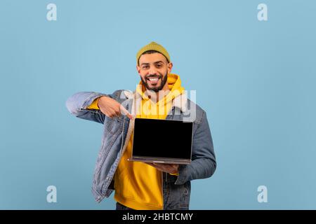 Ottimo sito web. L'uomo arabo eccitato mostra il computer portatile schermo vuoto e sorride alla fotocamera, sfondo blu, mockup Foto Stock