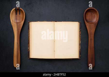 Apri un libro di cucina antico con pagine bianche e due cucchiai di legno, vista dall'alto, layout