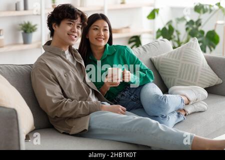 Buon fine settimana insieme. Giovane coppia asiatica affettuosa che beve caffè e coccola su un comodo divano a casa Foto Stock