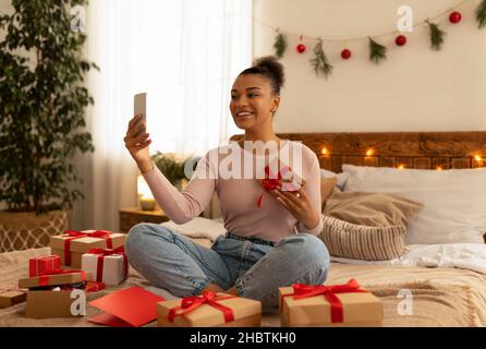 Festa di Natale online. Eccitata signora nera usando il cellulare, prendendo selfie o facendo videochiamata, celebrando le feste di Natale a casa Foto Stock