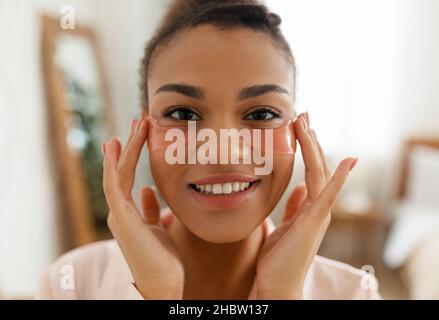 Attraente donna nera applicazione anti-affaticamento sotto la maschera degli occhi, cura della pelle, donna felice che tocca i cerotti sotto gli occhi, primo piano Foto Stock