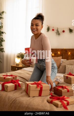 Eccitata donna afroamericana che tiene regali in mano e sorridente mentre si siede a letto circondato da regali di Natale Foto Stock