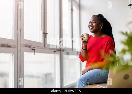 Giovane donna con tazza di caffè guardando fuori della finestra Foto Stock