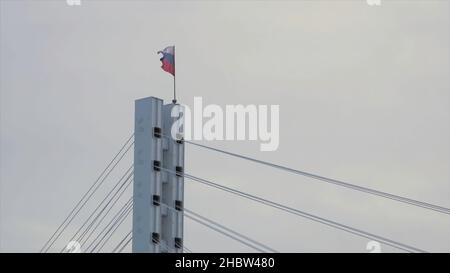 Bandiera della Federazione russa che sbatte nel vento sulla cima del ponte. Bandiera russa sulla cima di un ponte su sfondo grigio e nuvoloso. Foto Stock