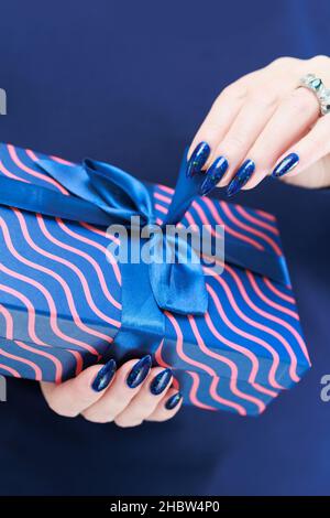 Le mani femminili con le unghie lunghe tengono una scatola del regalo nei colori blu Foto Stock