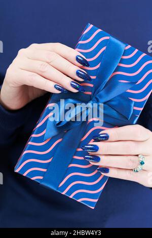 Le mani femminili con le unghie lunghe tengono una scatola del regalo nei colori blu Foto Stock