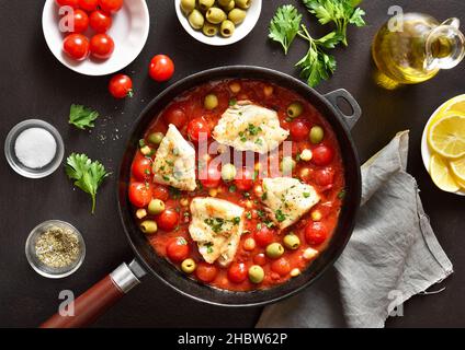 Stufato di merluzzo con ceci, pomodori ciliegini e olive in padella su sfondo di pietra scura. Vista dall'alto, piatto Foto Stock