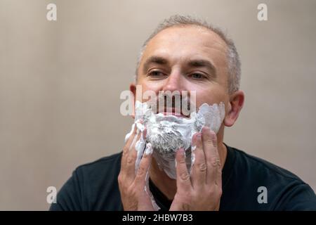 Ritratto di bel bianco-capelli beared uomo di mezza età applicando schiuma da barba per rifinire la barba Foto Stock