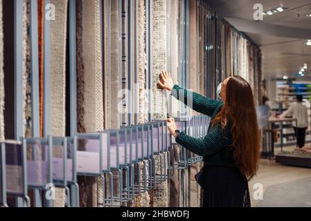 Donna giovane messa a fuoco in maschera medica scelta tappeto per la casa, facendo shopping durante covid19 epidemia, in piedi di fronte al tappeto stand guardando in su alla varietà di rivestimenti del pavimento in mostra Foto Stock